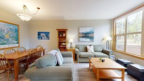 A living room with a sofa, a coffee table, a dining table with chairs, a bookshelf, and a large window. - Spacious 3BR Condo | Private Hot Tub & Morrisey Views (Sun Peaks)