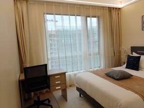 Et hotelværelse med en seng, et skrivebord med stol, et vindue med lette gardiner og udsigt til en bygning med stillads.
