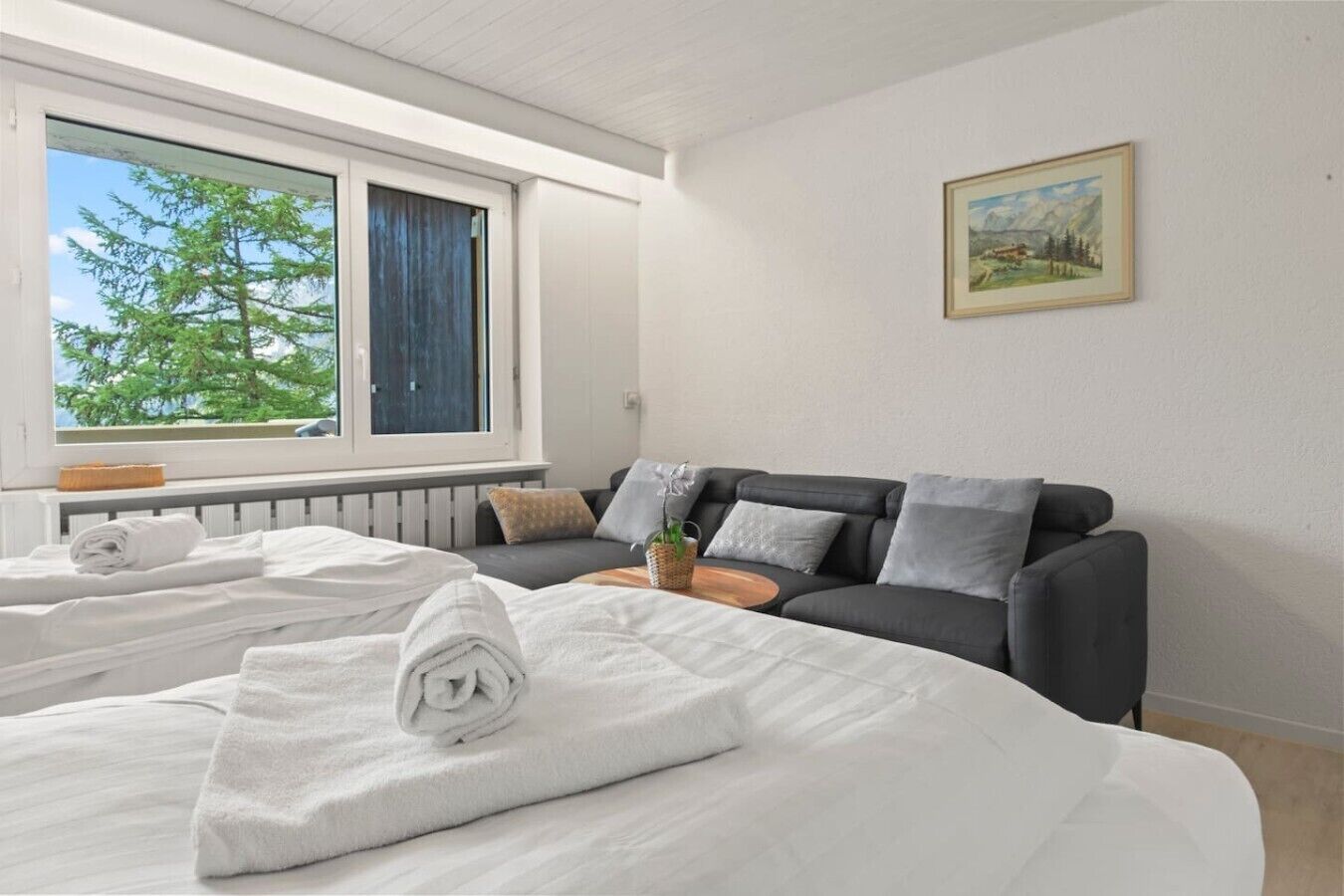 Ein Hotelzimmer mit Bett, Sofa, Fenster mit Blick auf Bäume und einem Gemälde an der Wand.
