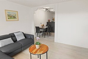 En moderne stue med en grå sofa, et rundt træsofabord og et spiseområde med bord og stole.