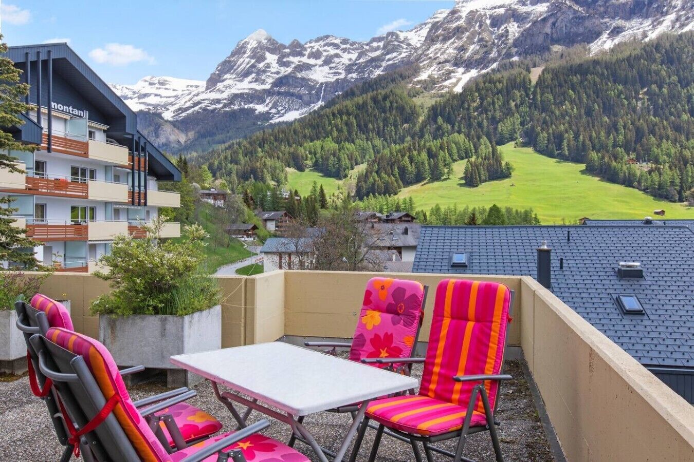 Un balcon avec des chaises et une table, d’où l’on peut admirer un paysage montagneux avec des bâtiments en arrière-plan.