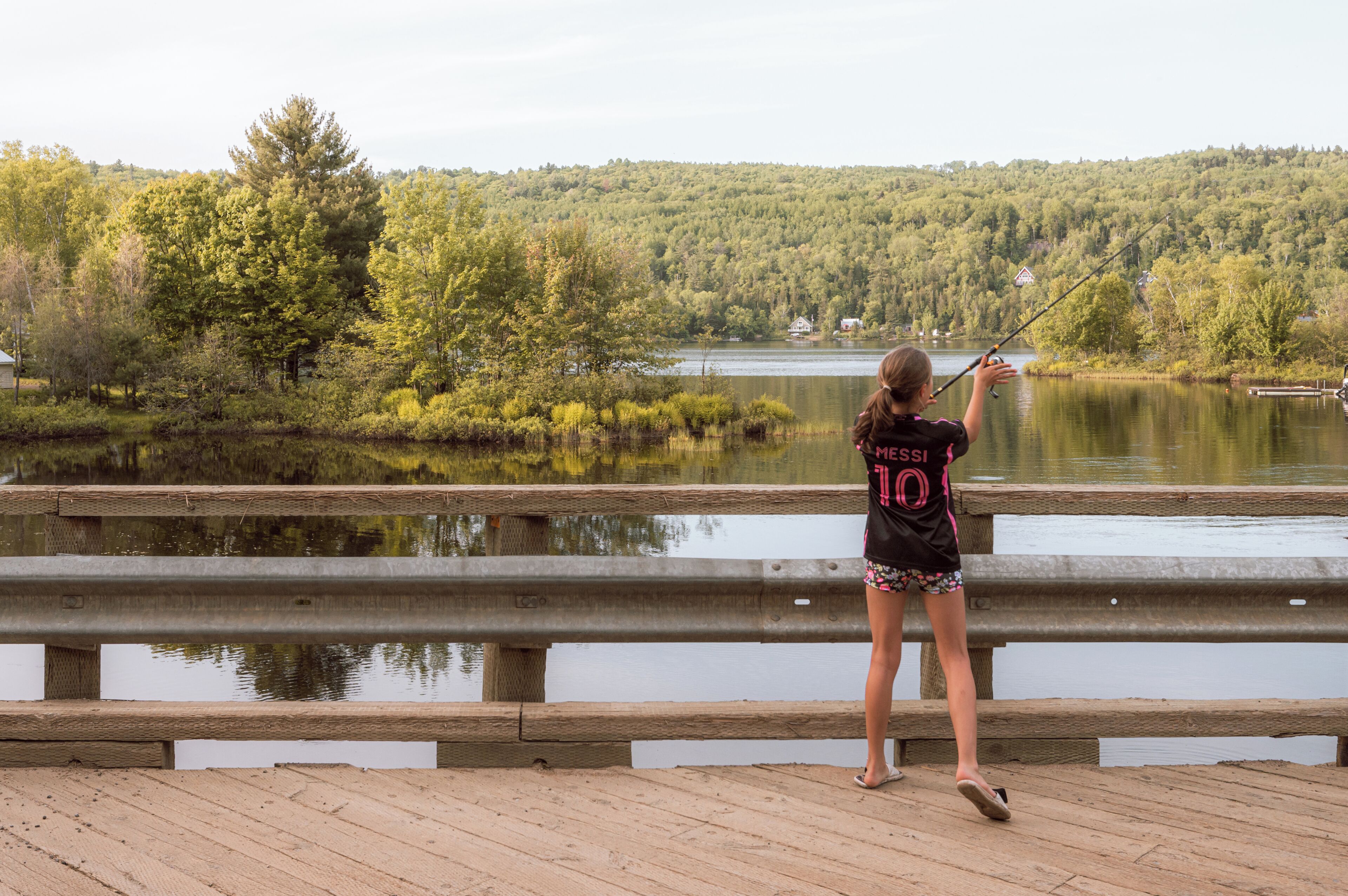 A person fishing on a wooden dock with a lake and forested hills in the background.