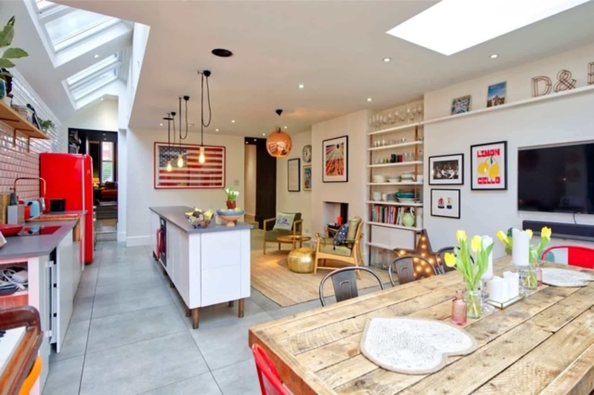A modern kitchen with a red refrigerator, a dining area with a wooden table, and a living room with a TV and bookshelves.