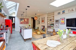 Eine moderne Küche mit einem roten Kühlschrank, ein Essbereich mit einem Holztisch und ein Wohnzimmer mit Fernseher und Bücherregalen.