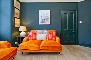 Ein Zimmer mit einer dunkelblauen Wand, einer grünen Tür und einem leuchtend orangefarbenen Sofa mit gemusterten Kissen.