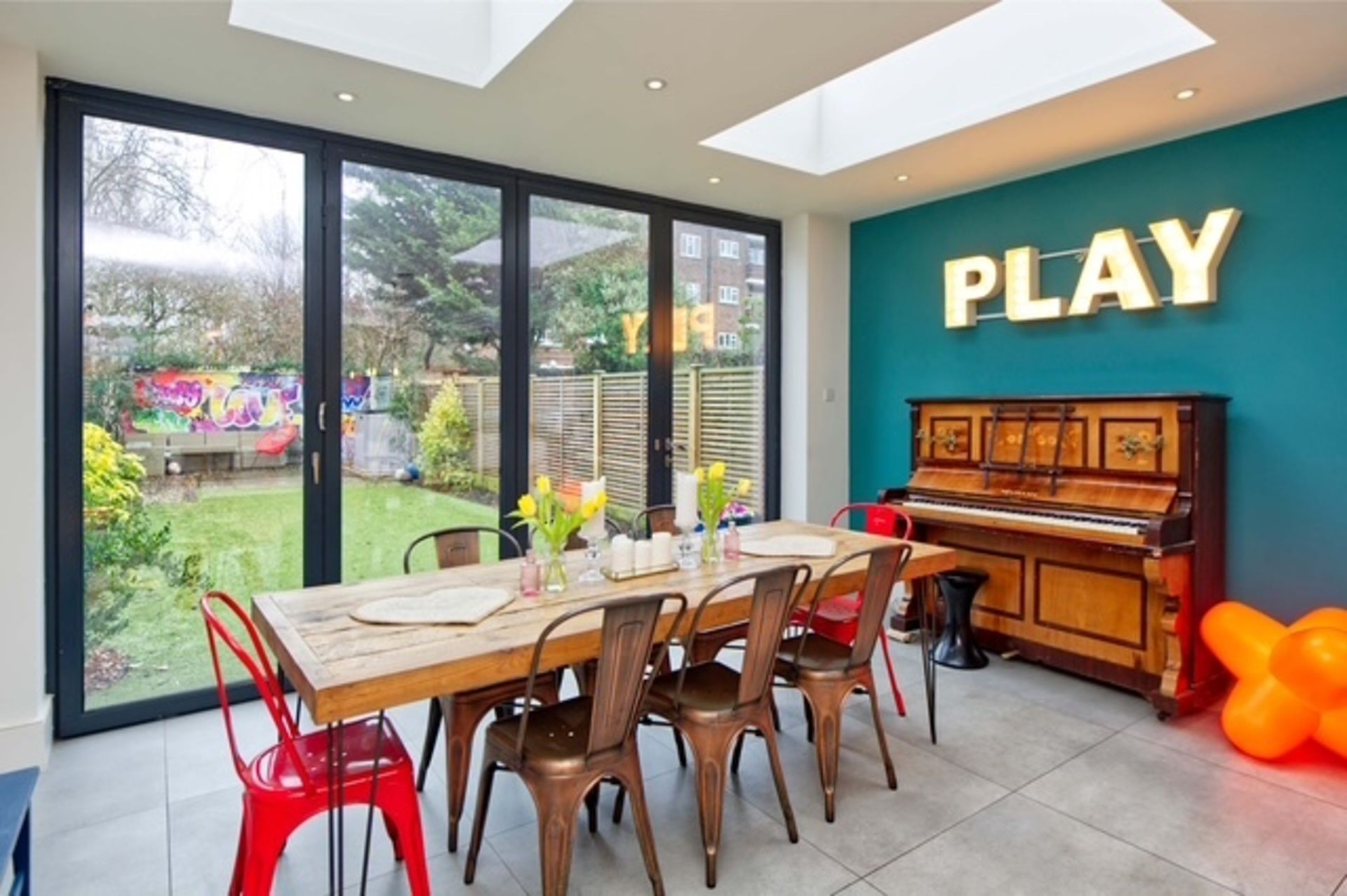 A dining area with a piano, a table with chairs, and a view through large glass doors to a garden.