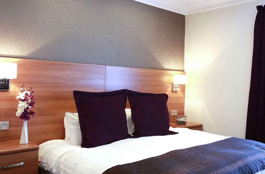 A hotel room with a wooden headboard, two bedside lamps, a vase with flowers, a bed with white linens, and a dark-colored throw blanket.