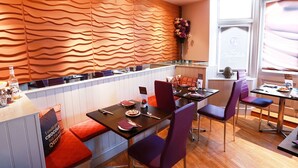Un restaurant au décor moderne, avec un mur aux motifs ondulés, un comptoir doté d’un distributeur de boissons et un espace salon agrémenté de chaises violettes et de coussins rouges.