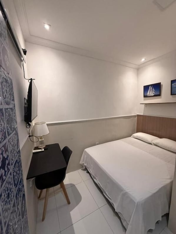 A hotel room with a bed, a desk with a chair, a lamp, and a wall-mounted TV.