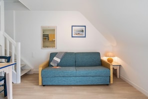 A room with a blue sofa, a small table with a lamp, a framed picture on the wall, and a kitchen visible through a mirror. - Scenic 6-person Duplex Retreat (Île-aux-Moines)