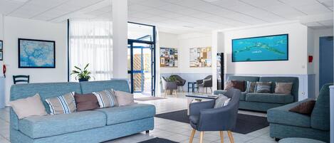 A modern living room with a blue sofa, a grey armchair, and a blue couch. There is a large window with curtains, a map on the wall, and a television displaying a scenic view.