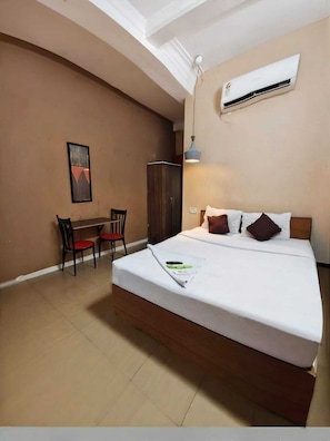 A bedroom with a bed, a desk, two chairs, an air conditioner, and a painting on the wall. - Nilay Guest House (Kolkata)