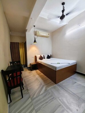 A compact room with a bed, a small table, a chair, a ceiling fan, and an air conditioning unit. - Nilay Guest House (Kolkata)