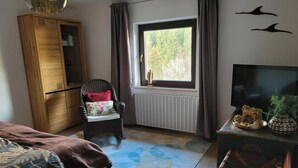 Ein Zimmer mit einem Bett, einem Holzschrank, einem Sessel mit Kissen, einem Heizkörper, einem Fenster mit Vorhängen, einem Fernseher und einer kleinen Zimmerpflanze.
