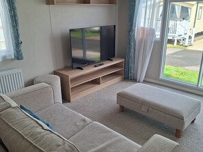 Una sala de estar con un televisor de pantalla plana sobre un soporte de madera, dos sofás y una ventana con cortinas.