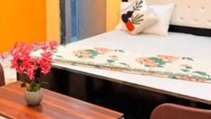 A bedroom with a bed, a bedside table, a chair, and a door. - Hotel Anuj Palace ll Near Ram Mandir - Comfortable AC Stay (Ayodhya)