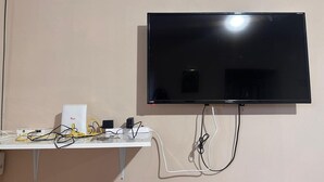 Un televisor de pantalla plana montado en la pared, con un estante blanco debajo, conectado a varios dispositivos electrónicos y cables.