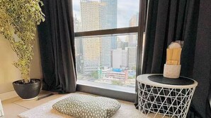 A modern room with a large window, a potted plant, a small round table, and a cushion on a beige rug. - BNEW! Cozy 1-BR Studio in High End Makati with Pool, Gym, Jacuzzi, Sauna (Makati)