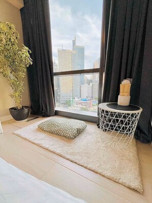 A modern room with a large window, a potted plant, a small round table, and a cushion on a beige rug. - BNEW! Cozy 1-BR Studio in High End Makati with Pool, Gym, Jacuzzi, Sauna (Makati)