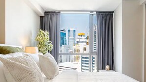 A hotel room with a large bed, a bedside table, a lamp, a potted plant, and a view of the cityscape through a large window. - BNEW! Cozy 1-BR Studio in High End Makati with Pool, Gym, Jacuzzi, Sauna (Makati)
