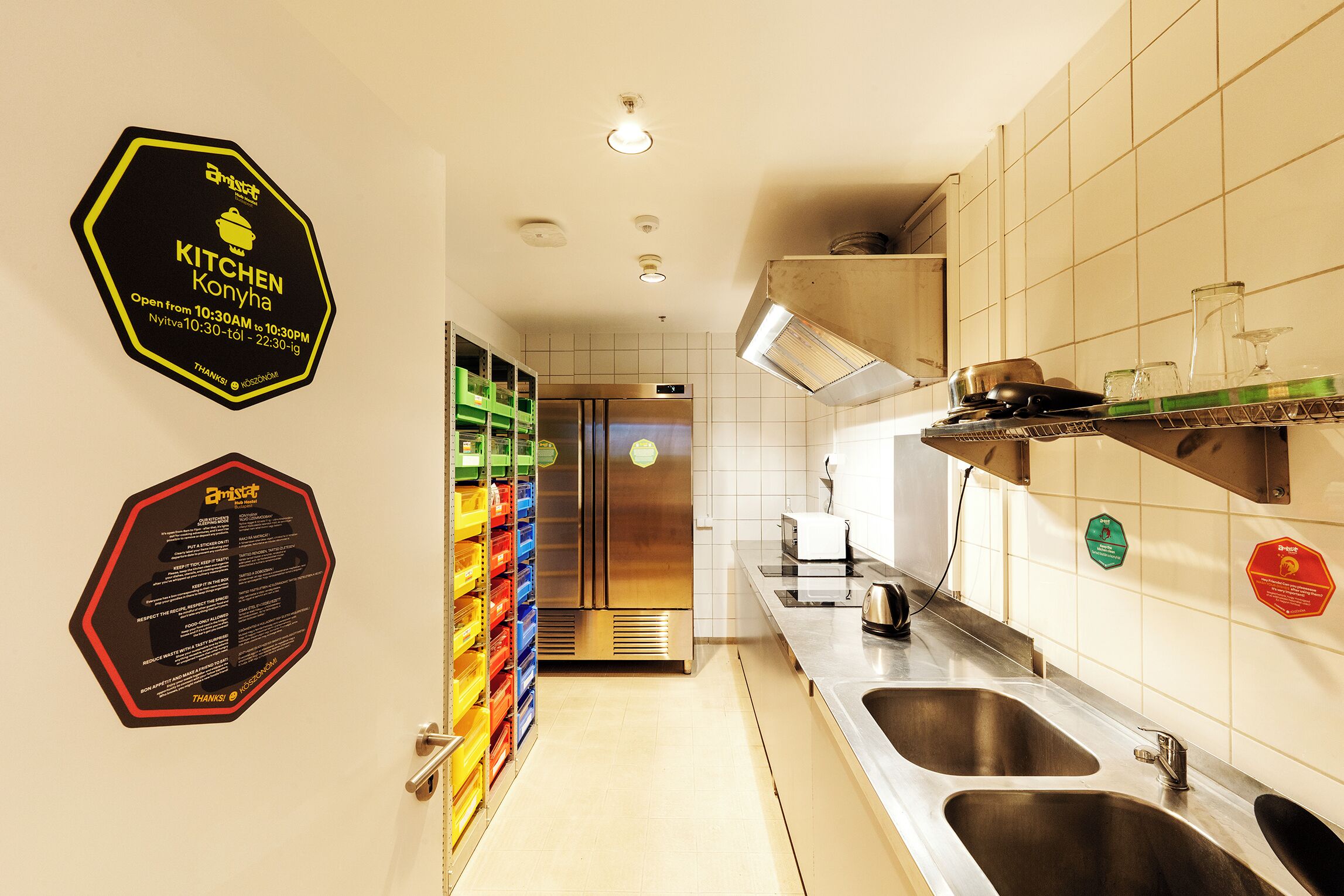 Una cocina moderna con electrodomésticos de acero inoxidable, un lavamanos y una encimera. Se observan estanterías y un cartel con el horario de atención de la cocina.