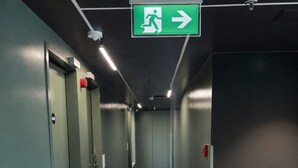 Un couloir avec des portes vertes, un panneau indiquant la direction de la sortie de secours et un feu rouge d’urgence.