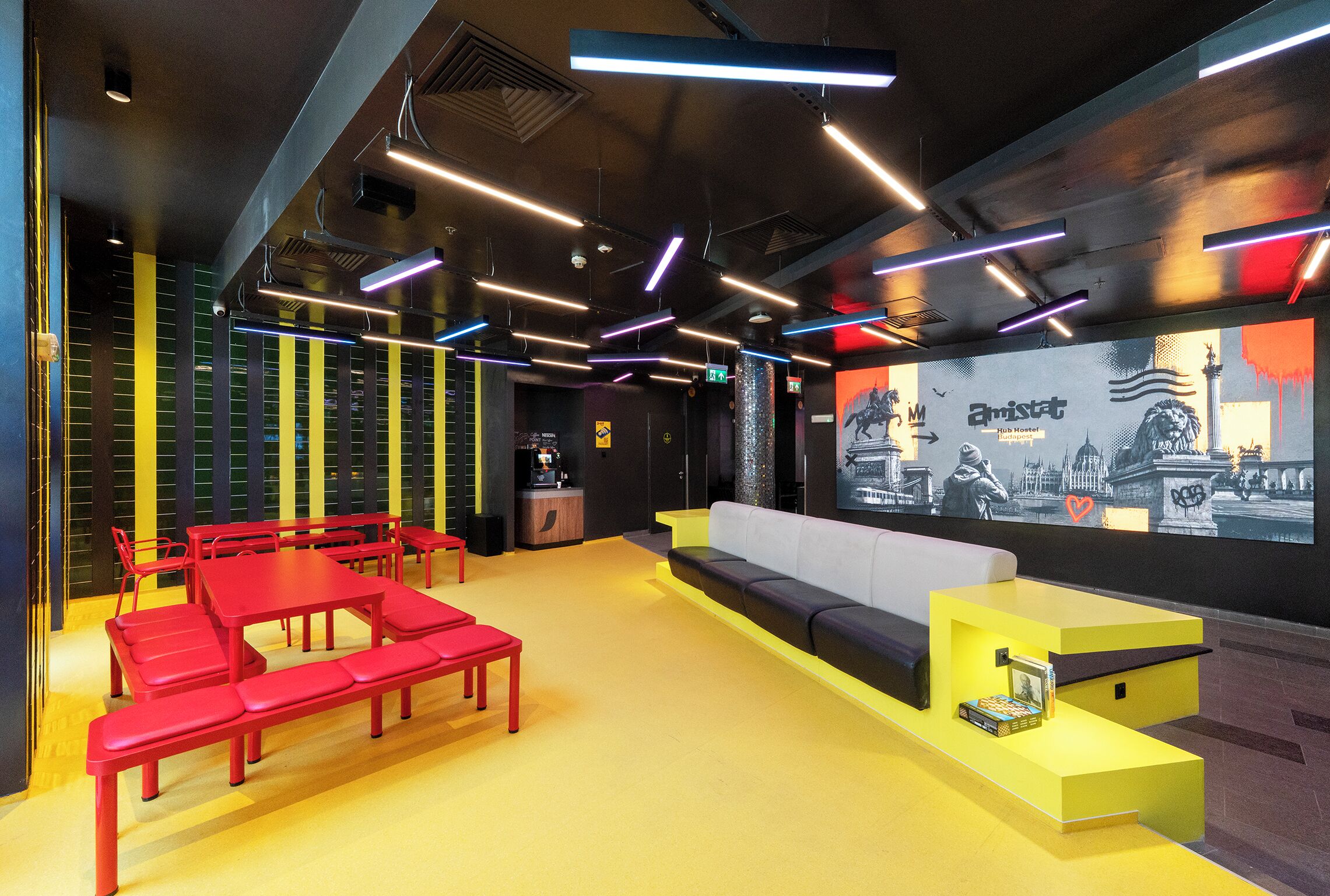 A modern lounge area with red seating, a yellow counter, and a large screen displaying a cityscape.
