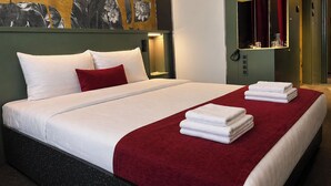 Une chambre d’hôtel avec un lit, un plaid rouge, des serviettes blanches, un miroir fixé au mur et une œuvre d’art aux motifs floraux.