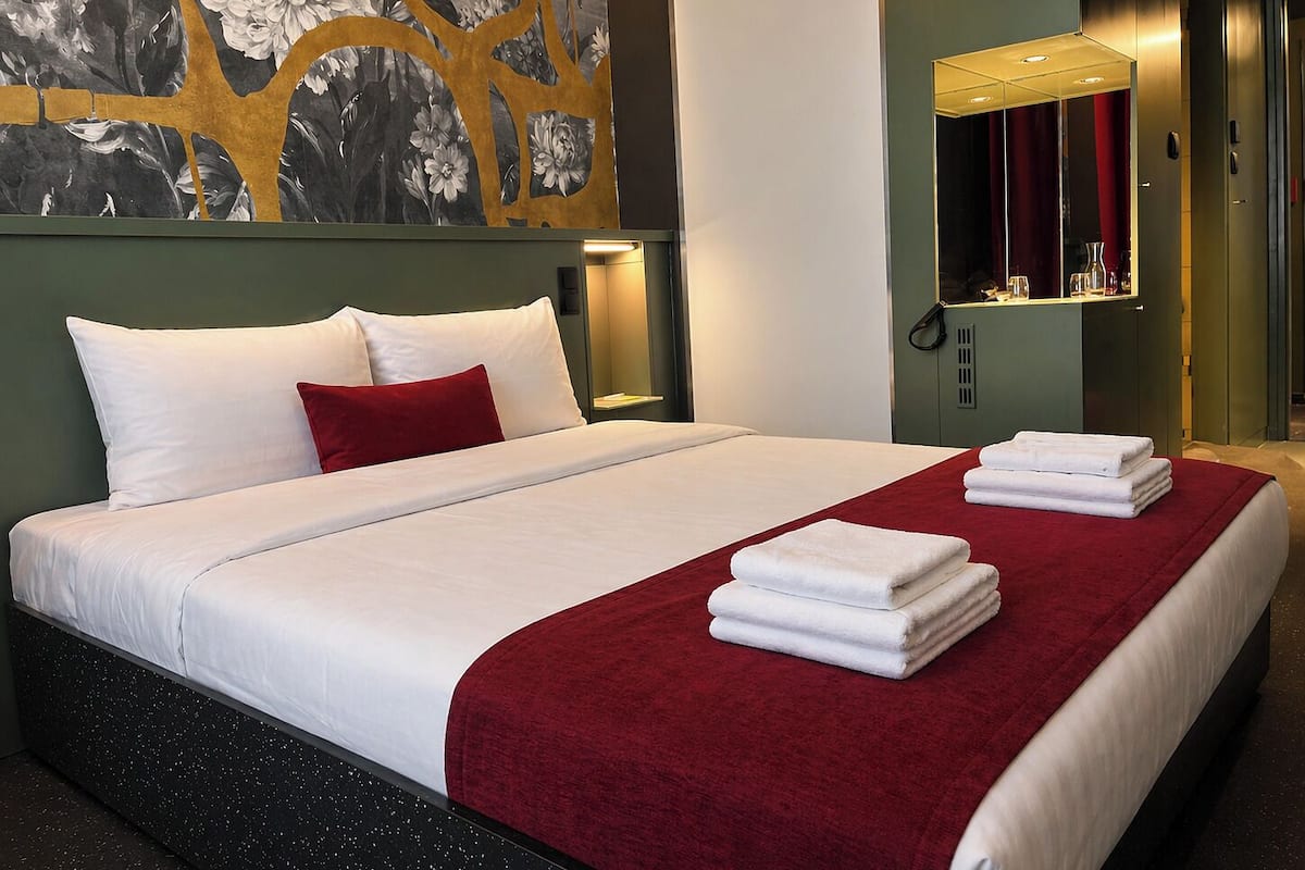 A hotel room with a bed, a red throw blanket, white towels, a wall-mounted mirror, and a floral artwork.