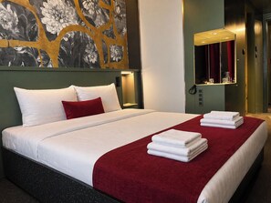 Hotelkamer met een bed, een rode plaid, witte handdoeken, een wandspiegel en een bloemrijk kunstwerk.