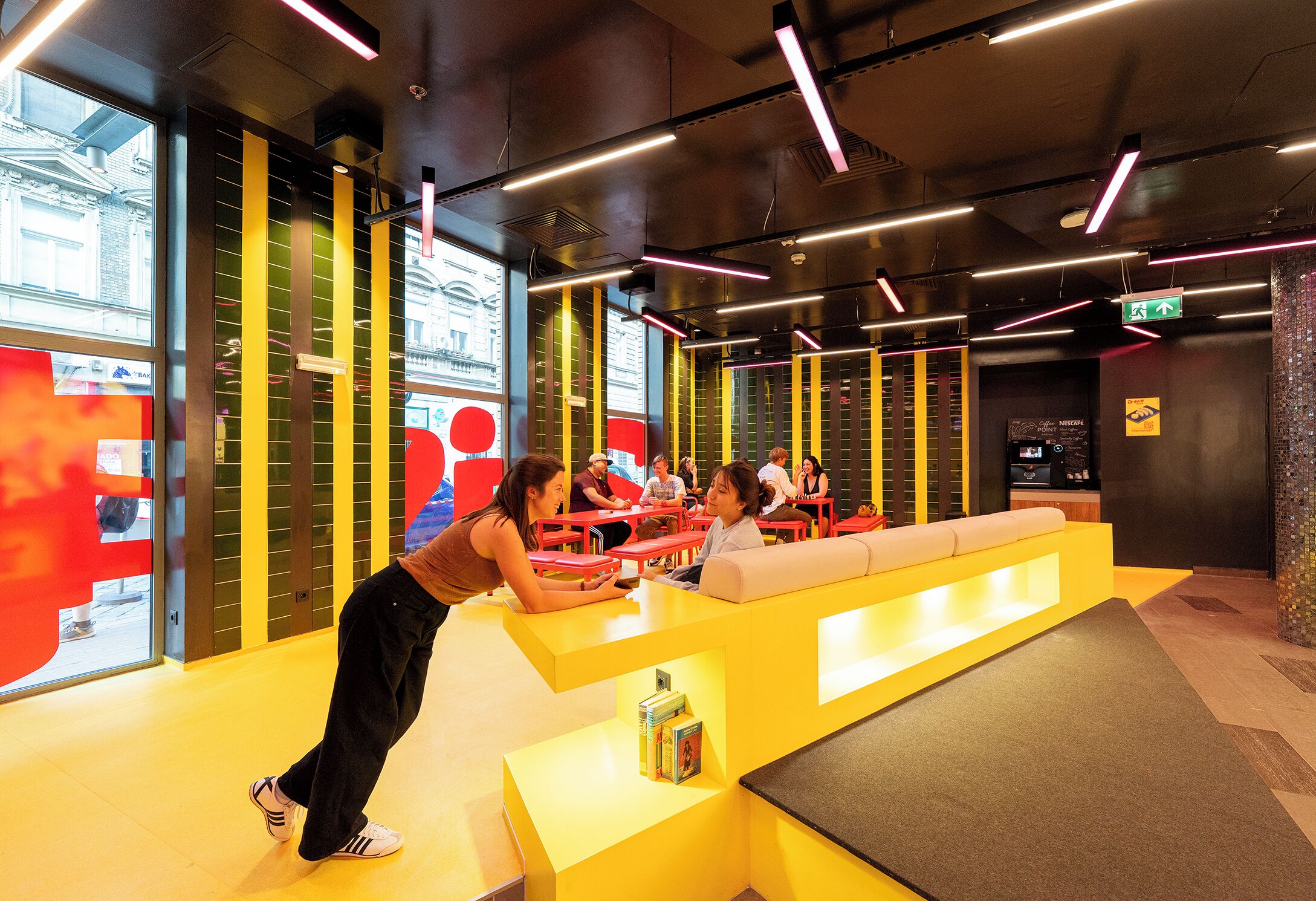 A modern indoor space with yellow and red seating areas, a reception desk, and a view of the outside through large windows.