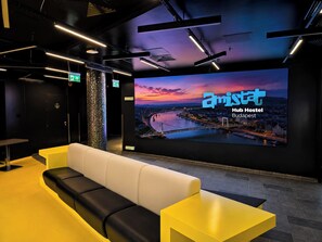 Un vestíbulo moderno con un sofá, una mesa amarilla y una pantalla grande que muestra 'amist Hub Hostel Budapest'.