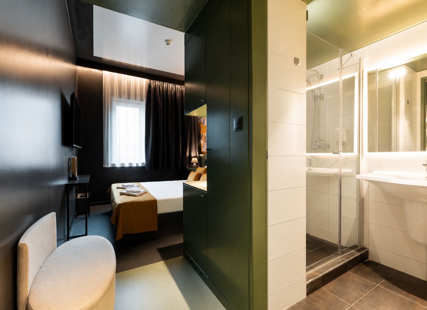 A hotel room with a bed, a desk, a chair, a window with curtains, and a bathroom with a sink and shower.