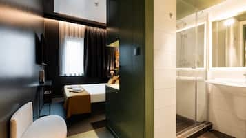A hotel room with a bed, a desk, a chair, a window with curtains, and a bathroom with a sink and shower.