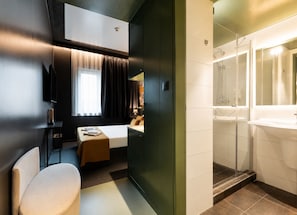 Habitación de hotel con cama, escritorio, silla, ventana con cortinas y baño con lavamanos y ducha.
