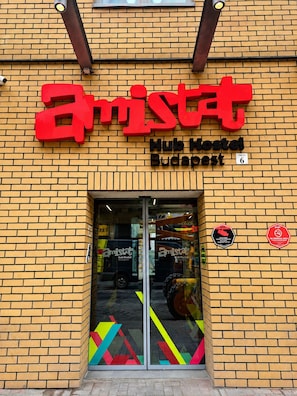 Een bord van Hub Hostel Boedapest met een rood en zwart logo, een bakstenen gevel en een glazen ingang.