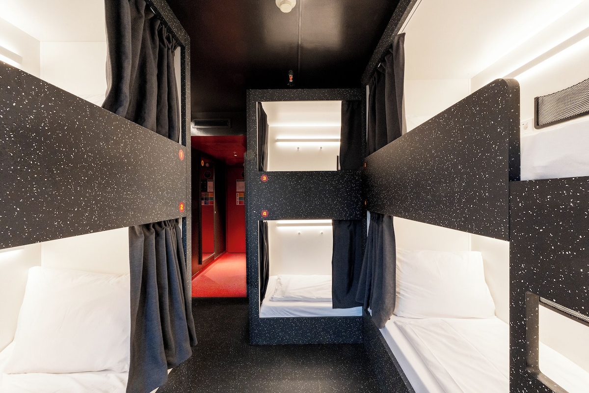 A narrow corridor with bunk beds on either side, leading to a red-lit room.