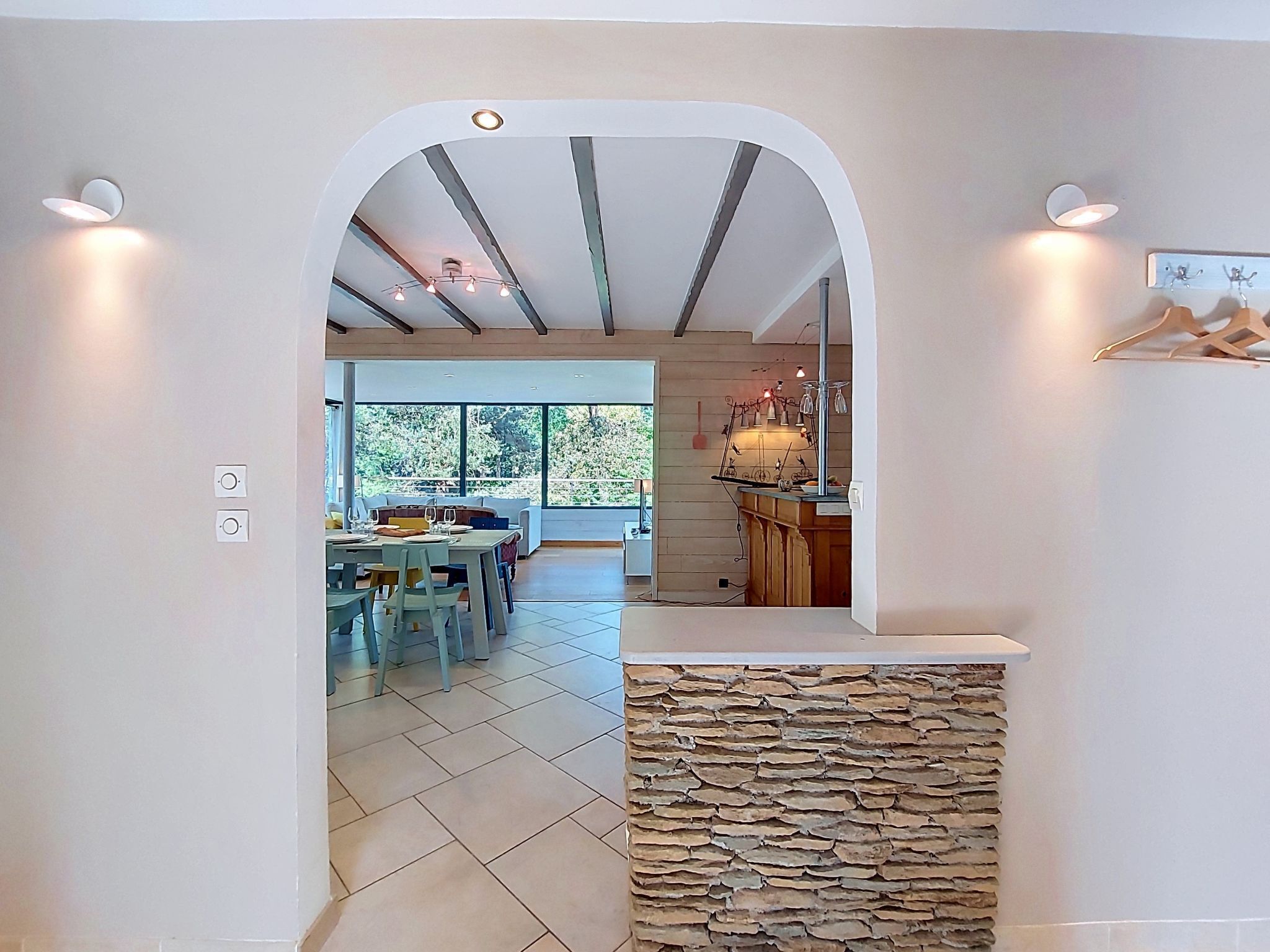 Un interior con pared de piedra, un área de comedor con sillas y una mesa, y una cocina con mostrador y lámparas colgantes.