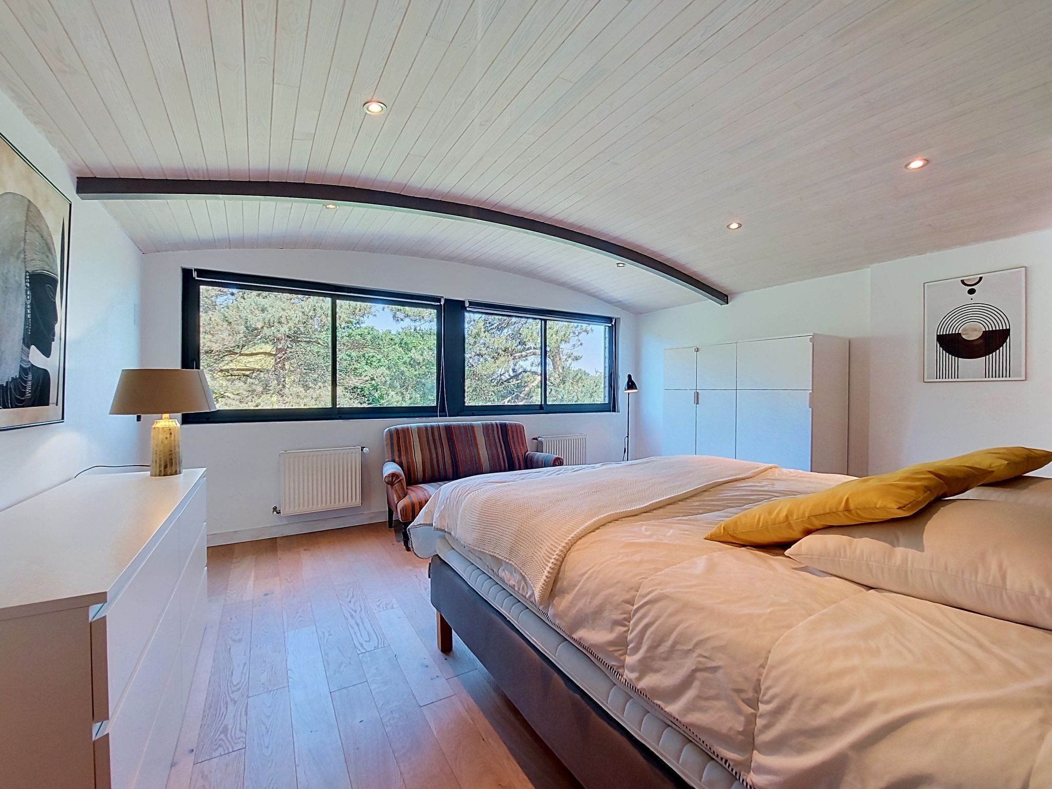Une chambre à coucher avec un plafond incurvé, une grande fenêtre, un lit avec un oreiller jaune, un radiateur et une lampe sur une table de chevet.