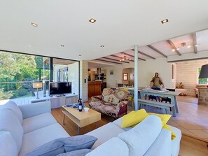 Ein modernes Wohnzimmer mit großem Fernseher, einer Couch, einem Essbereich und einer durchgehenden Tür erkennbaren Küche.