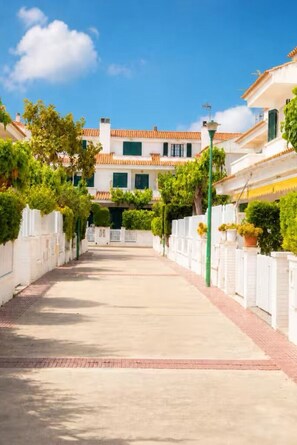 Una calle que lleva a un edificio con techo de tejas, paredes blancas y contraventanas verdes.