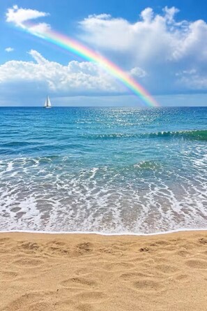 A beach with clear water, a sailboat, and a rainbow.