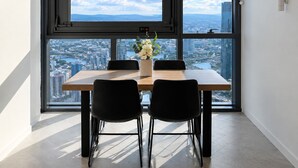 A dining area with a table and chairs, a vase with flowers, and a large window with a city view. - 63 Floors Up & A Million-Dollar View 2BR w/ Carpark (Brisbane City)