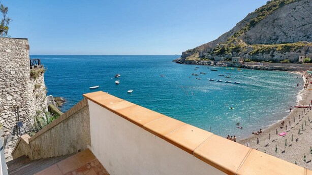 Vista costera con una escalera de piedra que conduce a un balcón con vista a una playa, barcos y una ladera rocosa.