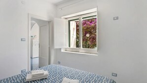 Un dormitorio con cama, ventana con vistas a flores, puerta que conduce a otra habitación y un interruptor de luz en la pared.
