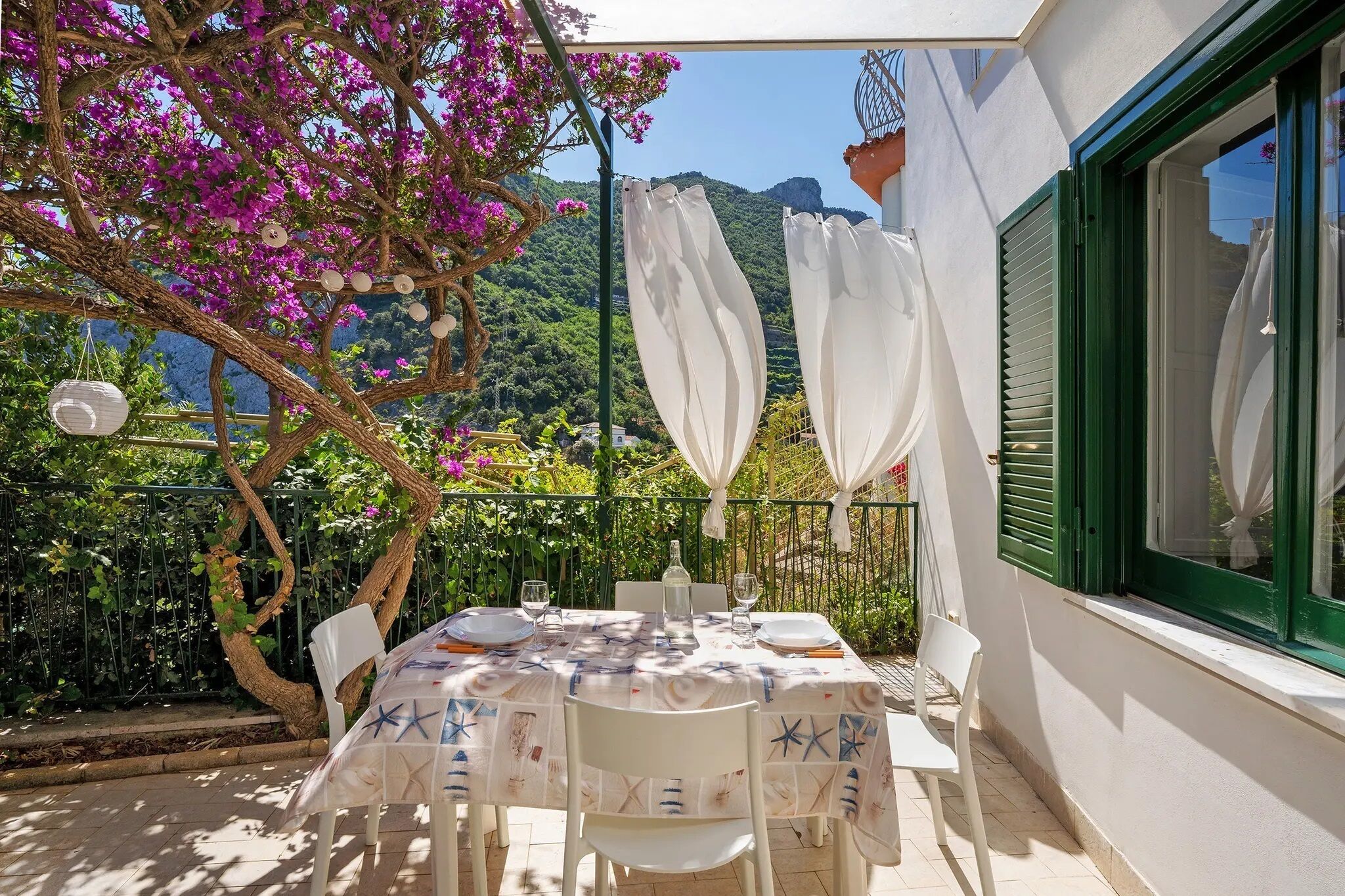 A dining table set outdoors with a view of mountains, a pergola with white curtains, and a tree with pink flowers.. BLUE APARTMENT AMALFI COAST with direct beach access