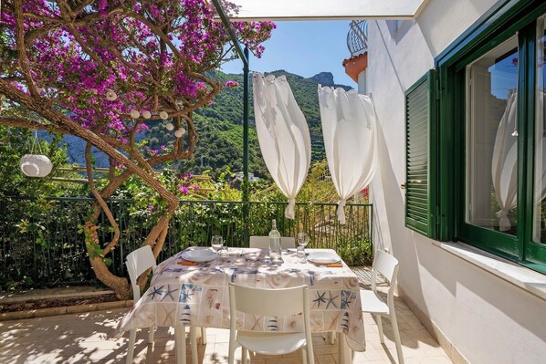 Ein Esstisch draußen mit Blick auf Berge, eine Pergola mit weißen Vorhängen und ein Baum mit rosa Blüten.