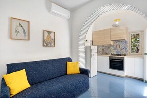 Ein Wohnzimmer mit einer Couch und zwei gerahmten Bildern an der Wand, ein rundbogiger Durchgang zu einer Küche mit Fliesenrückwand und einem Backofen.