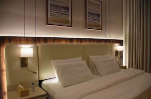 Una habitación de hotel con una cama, dos lámparas de noche, una mesita de noche con una caja de pañuelos y una botella de agua, y dos cuadros en la pared.
