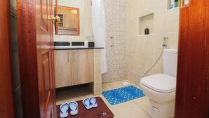 A bathroom with a toilet, sink, shower, and a pair of flip-flops on a mat. - Studio in Nairobi South B (Nairobi)